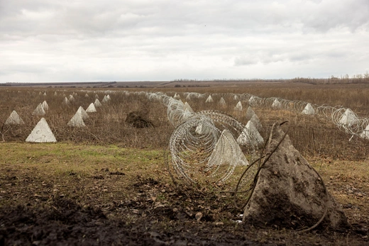 Rows of barbed wire and pyramidal anti-tank obstacles in a grassy field.