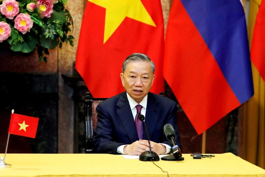 Vietnam's president wears a blue suit and purple tie while sitting at a desk with a microphone and two flags are behind him.