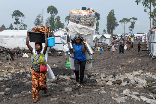 Refugees carry belongings, including a mattress and other supplies across a refugee camp.