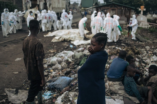 Civilians look on as workers in white hazmat suits collect and bury bodies in a dirt ground. 