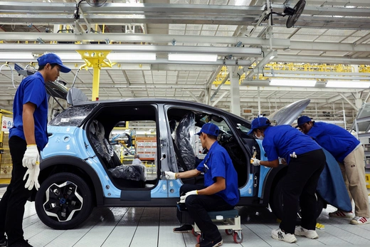 Workers wearing blue shirts and hats and black pants surround a car as they work on assembling it in a factory.