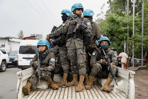 Armed UN peacekeepers in blue helmets stand and crouch on the bed of a truck.