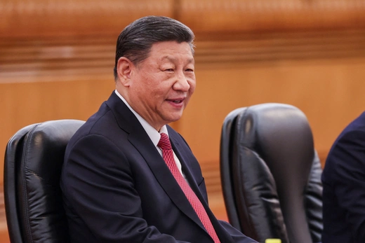 Chinese President Xi sits in a leather office chair in a wood-paneled room wearing a black suit and red tie.