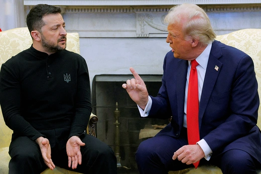 President Donald Trump is seated on a golden chair. He is pointing his finger in the air in front of Ukrainian President Volodymyr Zelenskyy who is also seated on a golden chair. He is wearing a black outfit and his open hands are gestured toward the floor.
