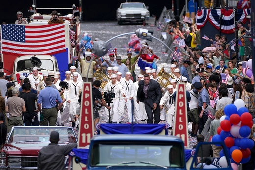 Actors in sailor costumes rush toward an arch on set surrounded by props including American flags, balloons, and "Apollo 11" signs adorning the arch.