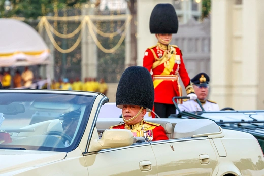 The Thai king sits in an open-top vehicle during a ceremony wearing a red coat wilth medals and a elongated black hat.