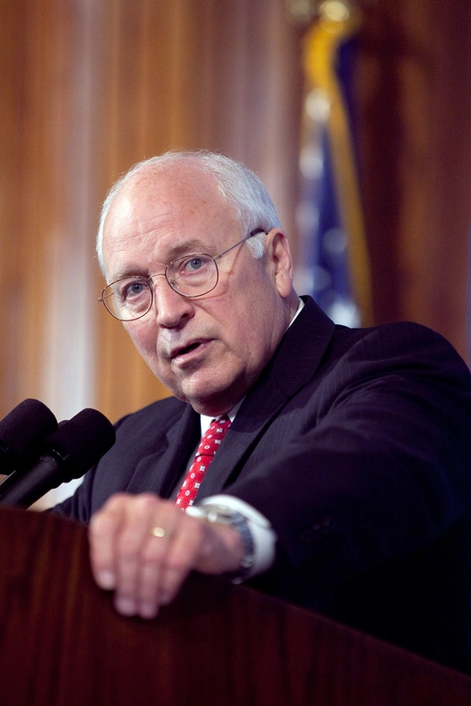 Dick Cheney stands at a podium with a microphone in front of him, staring to the left into the camera.