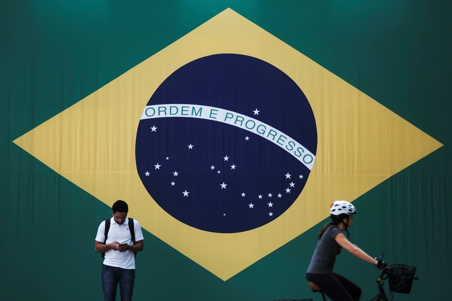 A man checks his mobile phone as a woman riding a bike passes next to a big Brazilian flag in Sao Paulo, Brazil June 28, 2018. 