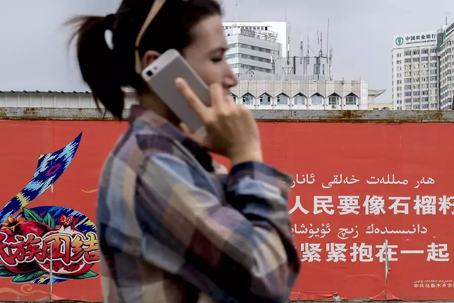 A young Uygur girl makes a phone call with her iPhone while walking past a propaganda wall.
