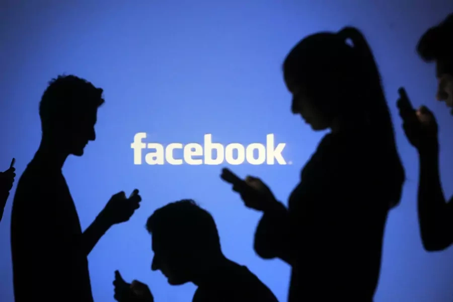 People are silhouetted as they pose with mobile devices in front of a screen projected with a Facebook logo