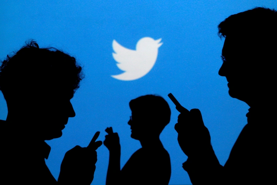 People holding mobile phones are silhouetted against a backdrop projected with the Twitter logo.