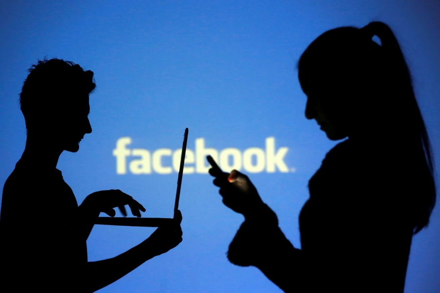 People are silhouetted as they pose with laptops in front of a screen projected with a Facebook logo.