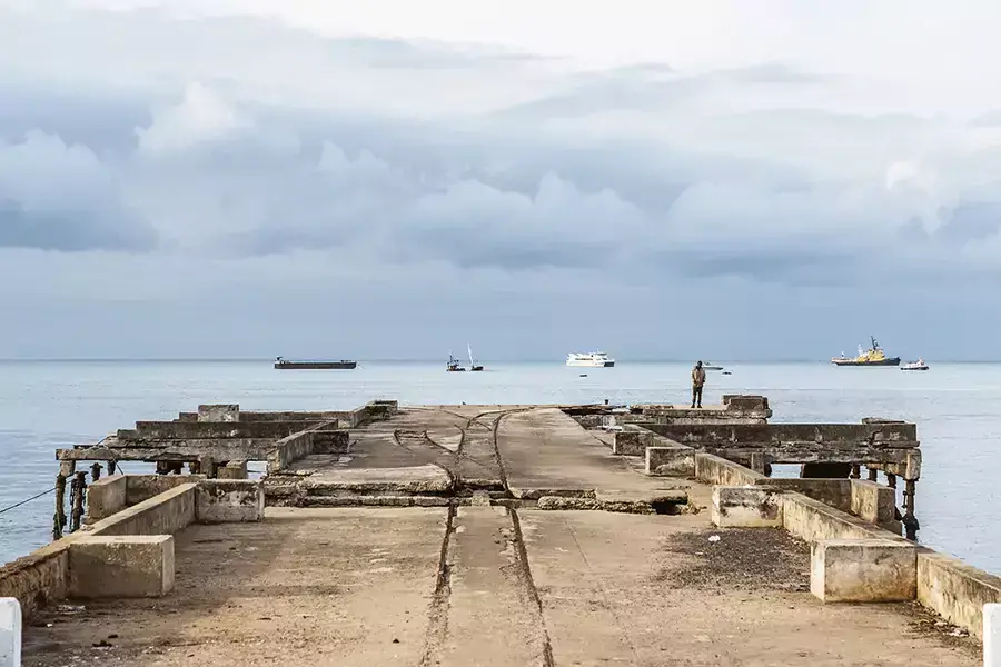 A lone man stands at the end of the old and broken pier watching the boats in the background in the bay of São Tomé city, São Tomé and Príncipe, September 16, 2021.